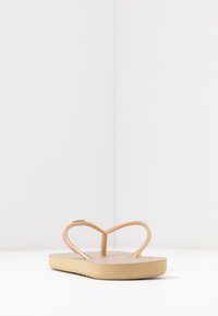 Tan flip-flop with a rubber toe strap in a heart shape, flat sole, and textured footbed, demonstrating a simple and functional design.