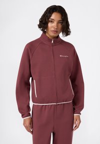 Burgundowa kurtka zapinana na zamek, wykonana z miękkiego materiału, z kołnierzem stojącym, kontrastowymi jasnymi wykończeniami i logo na lewej piersi. Posiada boczne kieszenie.