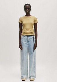 Tan fitted t-shirt, lichte blauwe wijde jeans met trekkoorddetails en beige instap schoenen. Model draagt zwarte zonnebril.