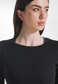 Czarna koszula z długim rękawem i zaokrąglonym dekoltem, wykonana z miękkiego materiału, z rękawami w stylu raglan. Nałożona na granatowy top na ramiączkach.