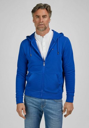 Man met grijs haar en baard, gekleed in een blauw vest met rits over een wit shirt en blauwe spijkerbroek, staand tegen een effen lichte achtergrond.