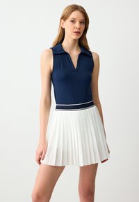 Navy mouwloze polo met een v-hals en een witte geplooide rok. Rok heeft een marineblauwe tailleband met een witte accentlijn. Glad oppervlak.