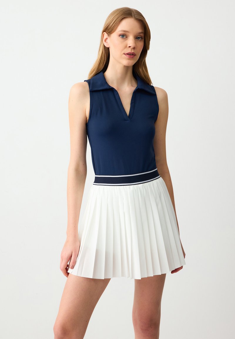 Navy mouwloze polo met een v-hals en een witte geplooide rok. Rok heeft een marineblauwe tailleband met een witte accentlijn. Glad oppervlak.