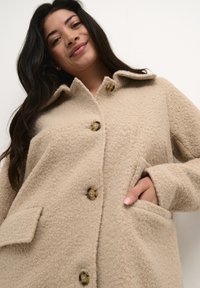 Beige, struktureret fleecejakke med krave, tre knapper og to frontlommer. Blødt materiale med et hyggeligt udseende.