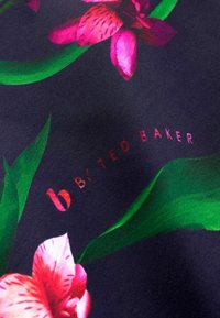 Donkere stof met levendige roze orchideeën en groene bladeren. De tekst luidt "b by TED BAKER" in het rood, dat opgaat in het bloemenpatroon.