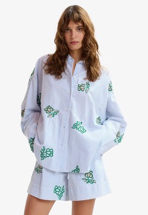 Donna con camicia larga a righe blu e bianche e pantaloncini con ricamo floreale verde, in piedi con le mani in tasca.