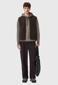 Męski strój składający się z brązowej, puchatej kamizelki bez rękawów nałożonej na szarą bluzę z kapturem, w połączeniu z czarnymi spodniami i czarnymi butami. Dodatki obejmują czarną torbę.