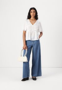 Blusa bianca a strati con dettagli in pizzo, abbinata a jeans a gamba larga a righe blu chiaro e una piccola borsa crema. Ballerine nere completano il look.