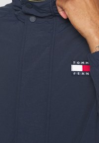 Marineblauwe windbreaker met een hoge kraag, voorzien van een logo-label in rood, wit en blauw, en zichtbare stikseldetails. Zachte, getextureerde stof.
