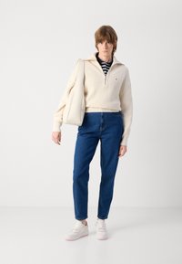 Lys beige strikkegenser med glidelås på kragen, blå rette jeans og hvite joggesko med rosa detaljer. Modellen holder en kremfarget håndveske.