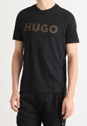 Homme portant un t-shirt noir uni à manches courtes avec le mot "HUGO" en grandes lettres marron foncé sur la poitrine.