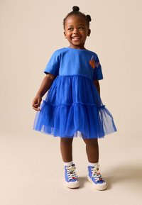 Petite fille souriante avec les cheveux en deux chignons porte une robe en tulle bleue et des baskets montantes bleues avec des écussons colorés.