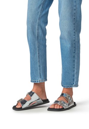 Silberne, flache Sandalen aus Metall mit zwei breiten Riemen, die dekorative Akzente aufweisen, auf einer schwarzen Sohle. Getragen zu knöchellangen blauen Jeans.