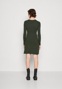 Abito lungo a maniche lunghe di colore verde scuro con silhouette aderente e orlo svasato, abbinato a stivaletti neri alla caviglia, esposto su uno sfondo neutro.