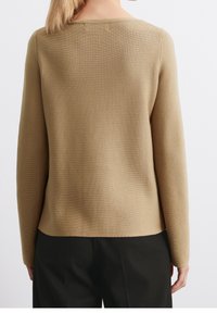 Femme portant un pull beige uni à manches longues et un pantalon noir, vue de dos sur un fond blanc.