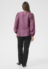 Donna di spalle che indossa una blusa lunga a maniche lucide viola con maniche a sbuffo e pantaloni neri a gamba dritta con tacchi neri.