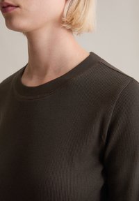 Pull côtelé vert olive foncé avec un col rond. Gros plan montrant le tissu texturé et un détail minimaliste de couture sur l'épaule.