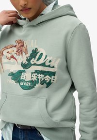 Persona che indossa un hoodie verde salvia con un design ricamato di uccello e onda, sovrapposto a una maglietta a righe e abbinato a jeans blu.