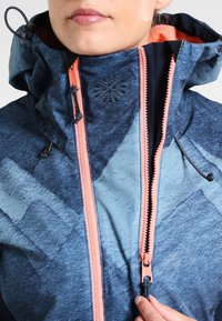 Giacca a motivi blu e grigia con collo alto, dotata di zip rosa e cordini regolabili sulla cappuccio e sull'orlo.