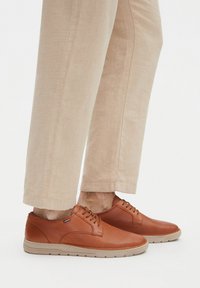 Hnědé kožené tenisky s texturovaným povrchem, kulatou špičkou a šněrovacím designem. Světle béžové kalhoty s rovný střih.