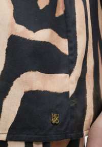 Μαύρο και μπεζ ύφασμα με αφηρημένο σχέδιο και μικρή χρυσή μεταλλική ετικέτα με ανάγλυφο το "HUGO" κοντά στο στρίφωμα.