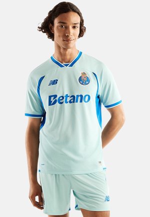 Junger Mann in hellblauem FC Porto Fußballtrikot mit Betano Sponsorlogo, vor einfachem weißen Hintergrund stehend.