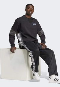 Černá mikina s šedými akcenty, se znakem Adidas. V kombinaci s černými teplákovými kalhotami, obě mají bílé pruhy. Obuto s bílými teniskami.