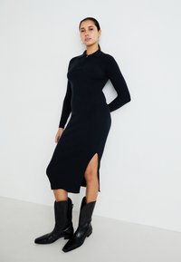 Black ribbed dress with long sleeves, a zip neckline, and side slits, paired with knee-high black boots with pointed toes and chunky heels.