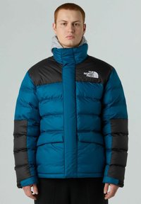 Giacca a vento blu e nera con un alto colletto, dotata di un logo sul petto, chiusura con zip frontale e tasche laterali. Tessuto liscio.
