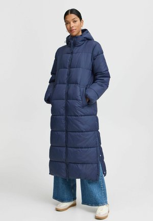 Navy long puffer coat with a hood, featuring quilted sections, side pockets, and snap closures at the hem, worn over wide jeans and sneakers.