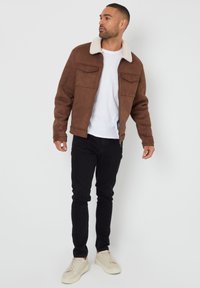 Brun ruskindsjakke med en hvid shearling-krave, to brystlommer og lynlås; parret med sorte bukser og lyse sko.
