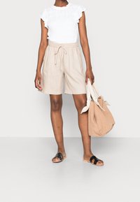 Shorts en lin beige avec une taille élastique, associés à un blouse blanche à volants et des sandales à enfiler noires. Tenant un sac fourre-tout beige.