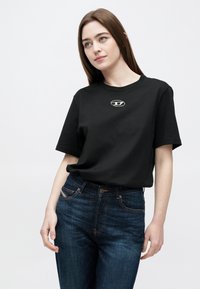 Μαύρο βαμβακερό t-shirt με στρογγυλή λαιμόκοψη και κοντά μανίκια, με μικρή λεπτομέρεια λογότυπου στο κέντρο. Φορεμένο με σκούρο τζιν παντελόνι.