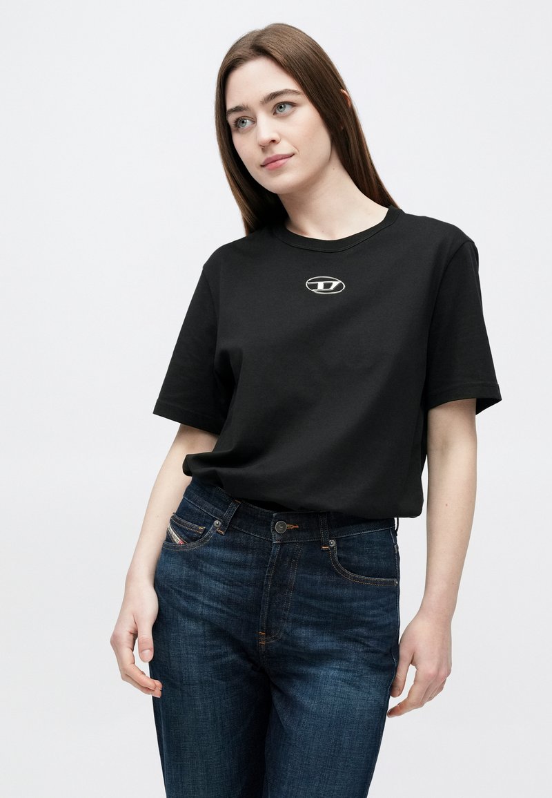 Camiseta negra de algodón con cuello redondo y mangas cortas, que presenta un pequeño detalle de logo en el centro. Combinada con pantalones vaqueros oscuros.