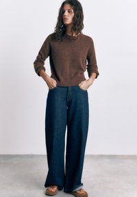 Žena valovite kose koja nosi smeđi džemper, široke tamnoplave traperice i smeđe sandale, stojeći s rukama u džepovima na jednobojnoj pozadini.