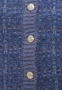 Πλεκτή μπλε ζακέτα με υφή, κάθετες ραβδώσεις και πέντε χρυσά κυκλικά κουμπιά κατά μήκος της εμπρός ραφής.