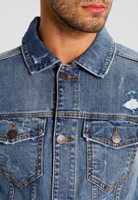 Džínová bunda s prošoupaným vzhledem, dvěma náprsními kapsami se stříbrnými knoflíky a klasickým límečkem ve středně modré barvě.