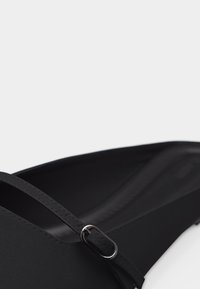 Μαύρο επίπεδο παπούτσι με λεπτό ρυθμιζόμενο λουράκι και ασημένια αγκράφα, κοντινή θέα στο λείο υλικό και τις λεπτομέρειες ραφής.