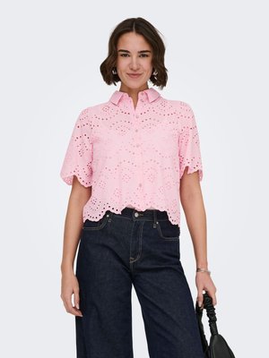 Donna che indossa una camicetta rosa con bottoni ad occhiello, maniche corte e orlo smerlato, abbinata a jeans scuri, con una borsa nera in mano.
