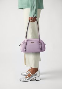 Bolso de mano morado con una textura suave, asas dobles en la parte superior y acentos plateados. Llevado con pantalones claros y zapatos plateados brillantes.