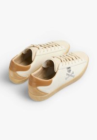 White leather sneakers with tan accents, textured heel, beige sole, and embroidered skull and crossbones design on the side.