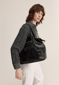 Černá kožená kabelka s hladkou texturou a jemným leskem. Má pevné rukojeti a zip, doplněná tlustě pleteným šedým svetrem a bílými kalhotami.