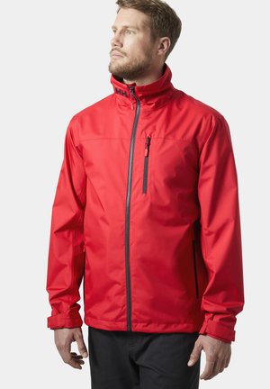 Mężczyzna w jaskrawoczerwonej kurtce na zamek z wysokim kołnierzem i zapinanym na zamek kieszonką na piersi, patrzący w lewo na jednolitym tle.