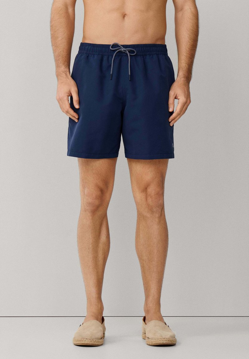 Homme debout portant un short de bain bleu marine avec cordon de serrage et des chaussures beige à enfiler, sur un fond neutre.