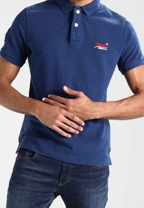 Homme portant un polo bleu et un jean, les mains jointes sur le ventre devant un fond clair uni.