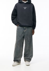 Ciemnoszara bluza z kapturem z przednią kieszenią, zestawiona z szerokimi, jasnoniebieskimi spodniami w paski i białymi tenisówkami. Prosty, swobodny fason.