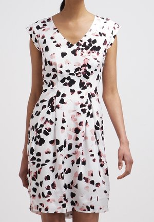 Femme portant une robe blanche à manches courtes avec des motifs abstraits tachetés noirs et roses, debout devant un fond uni.
