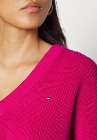 Rosa Strickpullover mit V-Ausschnitt und gerippter Textur, ausgestattet mit einem kleinen Logo-Patch auf der linken Seite nahe dem Ausschnitt.