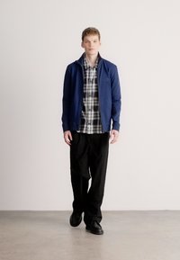 Navy zilā rāvējslēdzēja jaka pār melnbaltu pleda kreklu, kombinēta ar vaļīgām melnām biksēm un tumšiem apaviem, stāvošs pret gaišu fonu.
