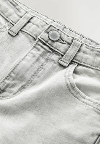 Gros plan sur un jean en denim gris clair montrant la taille, un bouton en métal avec texte, des passants de ceinture, une poche avant et des coutures détaillées.